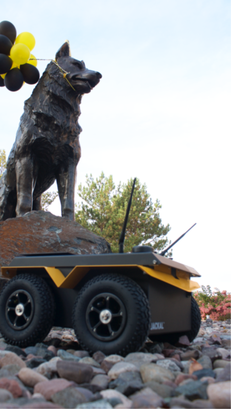 A Clearpath Jackal robot in front of the Husky statue on the Michigan Tech campus